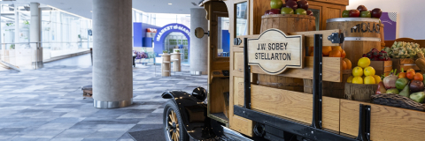 This image shows, Wooden cart with the sign "J.W. Sobey Stellarton" displaying various fruits and vegetables in barrels and baskets.