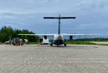 A small plane is parked on a tarmac against a backdrop of cloudy skies and green trees. Orange cones are placed around the area, suggesting it is an active work zone.