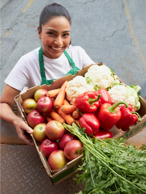 This banner image shows a woman holding a box of fresh vegetables and smiling while her picture is being taken.