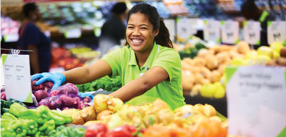 A smiling grocery store worker arranges fresh produce, including peppers and onions, on a display inside a market.