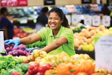 A smiling grocery store worker arranges fresh produce, including peppers and onions, on a display inside a market.