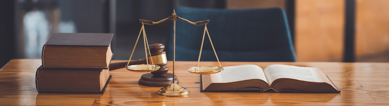 This image showing, a brass balance scale sits on a wooden table beside an open book and two stacked closed books. A wooden gavel rests behind the balance scale, near the corner of the table.