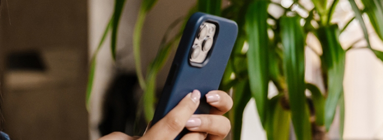 A person is holding a smartphone with a blue case, standing next to a green potted plant.